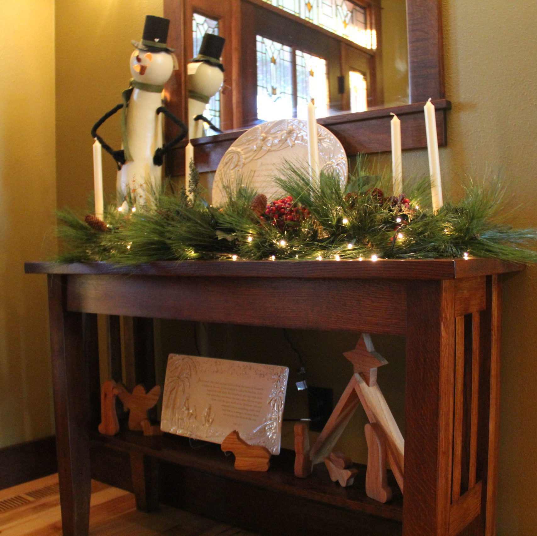 Front hall table complete with greenery, lights, tall pillar candles, a Meadowbrooke Gourd, a couple platters about the Christmas story and a handmade Nativity