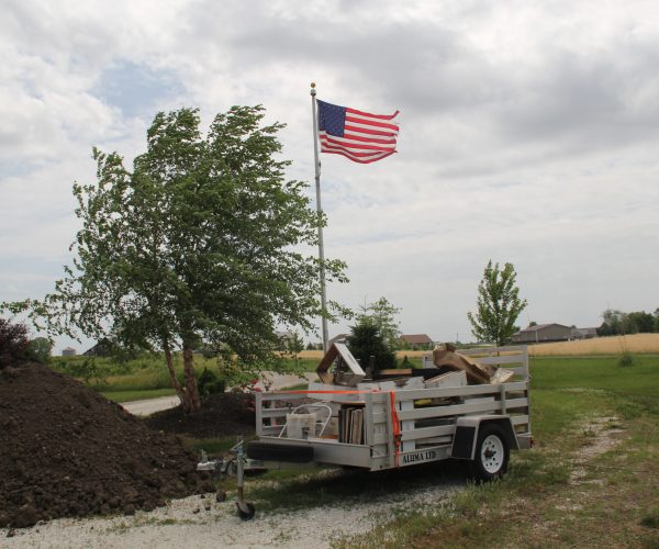 And here we show more work to do. Notice the trailer, instead of full of snow, it's full of bee equipment. A pile of dirt to backfill around the foundation. And our Ryan flag. She's taking it all as he is away and still rippling beautifully. Thank goodness, I need her strength some days.